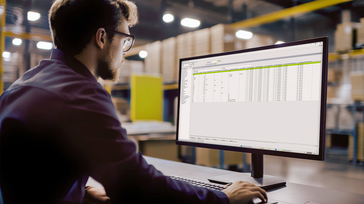 Ein Mann sitz am Schreibtisch im Lager und schaut sich Tabellen in der Software PSIwms am Monitor an.