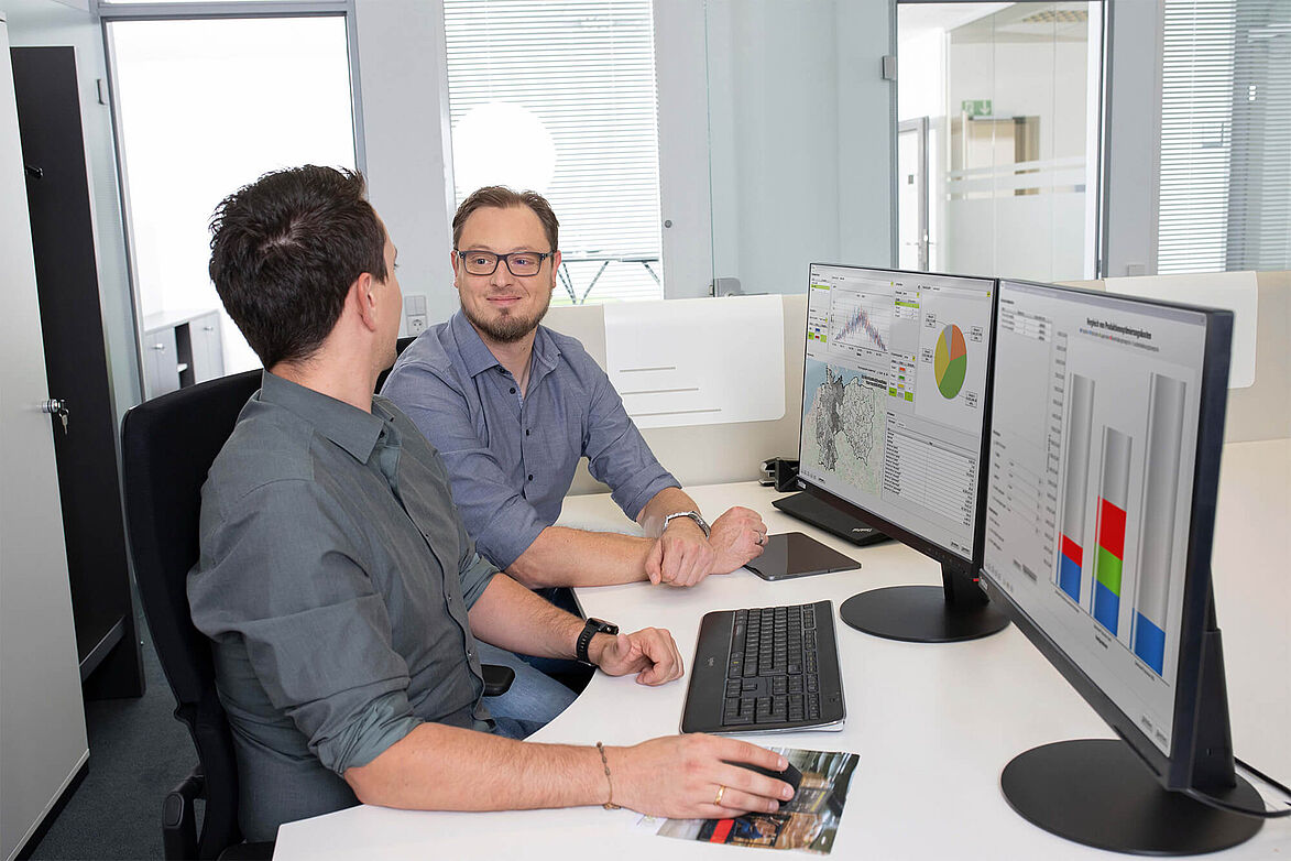 Zwei Personen auf dem Bild sitzen am Schreibtisch und unterhalten sich über die Software am Monitor.